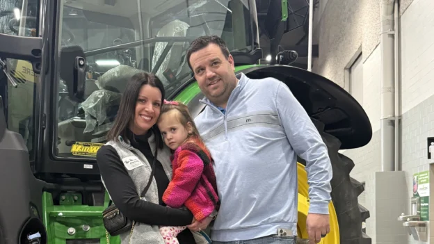 A man, woman, and a child pose in front of a large John Deere tractor at the Iowa Ag Expo.